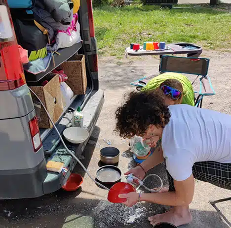 vanlifeuse faisant la vaiselle à l'arrière de son van aménagé grâce à une douchette